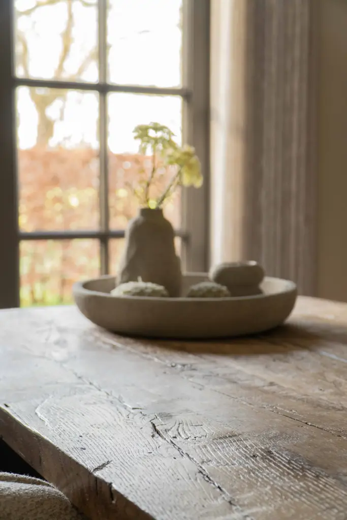 Close-up of a rustic reclaimed oak dining table with a naturally weathered surface beside a large window.